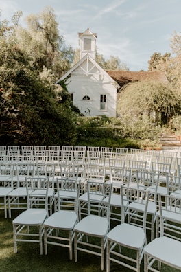 A quaint, white chapel surrounded by lush greenery, with a collection of white chairs arranged on a patch of grass in the foreground. The chapel has a small bell tower and features simple, classic architectural details. The setting appears serene and prepared for an outdoor event, possibly a wedding.