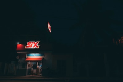 A small convenience store is seen at night, illuminated by its bright red and white signage reading 'SIX'. The store interior is dimly visible through the open front, displaying shelves of products. Surrounding the store, the environment is dark, with minimal lighting and a palm tree shadowed in the background.