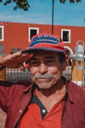 An elderly man with a mustache wearing a red and blue cap is saluting with his right hand. He is dressed in a red and black button-up shirt, standing in front of a red building with white-trimmed windows and a wrought iron fence. The sky is clear and blue, and part of a tree is visible in the upper left corner.