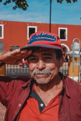 An elderly man with a mustache wearing a red and blue cap is saluting with his right hand. He is dressed in a red and black button-up shirt, standing in front of a red building with white-trimmed windows and a wrought iron fence. The sky is clear and blue, and part of a tree is visible in the upper left corner.