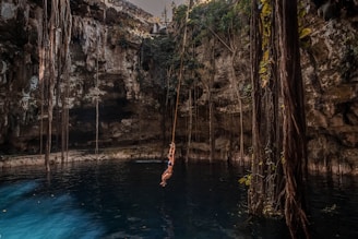 A person in a swimsuit swings on a rope over a deep blue cenote pool surrounded by high rocky walls with long roots and vegetation hanging down. The setting is serene with natural light filtering from above.