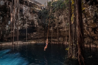 A person in a swimsuit swings on a rope over a deep blue cenote pool surrounded by high rocky walls with long roots and vegetation hanging down. The setting is serene with natural light filtering from above.