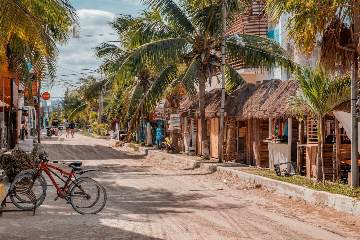 Bicycle parked by palm trees on a sand street lined with thatched shops, the slow pace that makes Holbox worth it for many visitors