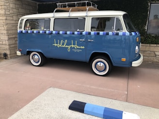 A vintage blue van with a white roof and decorative elements is parked on a paved area. The side of the van features the words 'Holiday House' in yellow script, suggesting it is affiliated with a specific location. The van has whitewall tires and a roof rack, with a window displaying stickers. The setting includes stone walls and greenery, indicating an outdoor urban or suburban environment.
