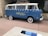 A vintage blue van with a white roof and decorative elements is parked on a paved area. The side of the van features the words 'Holiday House' in yellow script, suggesting it is affiliated with a specific location. The van has whitewall tires and a roof rack, with a window displaying stickers. The setting includes stone walls and greenery, indicating an outdoor urban or suburban environment.