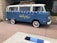 A vintage blue van with a white roof and decorative elements is parked on a paved area. The side of the van features the words 'Holiday House' in yellow script, suggesting it is affiliated with a specific location. The van has whitewall tires and a roof rack, with a window displaying stickers. The setting includes stone walls and greenery, indicating an outdoor urban or suburban environment.