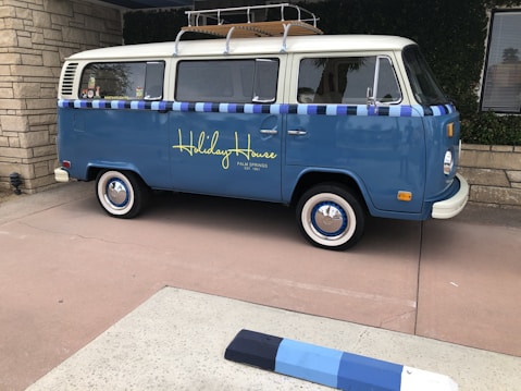 A vintage blue van with a white roof and decorative elements is parked on a paved area. The side of the van features the words 'Holiday House' in yellow script, suggesting it is affiliated with a specific location. The van has whitewall tires and a roof rack, with a window displaying stickers. The setting includes stone walls and greenery, indicating an outdoor urban or suburban environment.