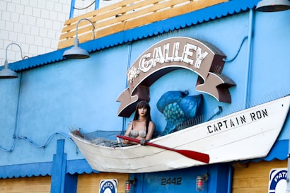 A mannequin dressed as a mermaid is sitting in a boat mounted on a wall outside. The wall is painted blue and features a sign that reads 'The Galley'. The boat is labeled 'Captain Ron', and a large blue fish sculpture is positioned behind the mannequin. The setting includes nautical elements and decorative lights.