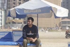 A man sits under a large beach umbrella branded with the word 'Berger' at a sandy area, possibly a beach or construction site. He wears a dark blue sweatshirt and olive pants. Behind him, there are buildings under construction, a vehicle, and some distant figures.