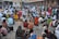 A vibrant gathering of people is taking place in what appears to be a religious or cultural setting. Many individuals are seated on the ground, surrounded by numerous small lit candles. The attendees are dressed in colorful traditional garments. The environment is bustling, with some people engaged in prayer or meditation, while others are arranging the candles into patterns or designs on the ground. An ancient stone pillar and ornate architecture are visible in the background.