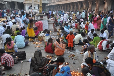 A vibrant gathering of people is taking place in what appears to be a religious or cultural setting. Many individuals are seated on the ground, surrounded by numerous small lit candles. The attendees are dressed in colorful traditional garments. The environment is bustling, with some people engaged in prayer or meditation, while others are arranging the candles into patterns or designs on the ground. An ancient stone pillar and ornate architecture are visible in the background.