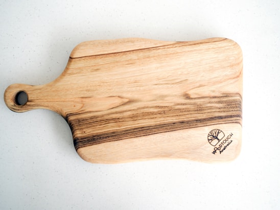 A wooden cutting board with natural wood grain patterns, highlighting darker and lighter streaks. The board has a handle with a hole for hanging. A logo with the text 'WOODTOUCH Australia' is engraved in one corner.