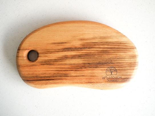 A wooden cutting board with a smooth, rounded shape and a large hole on one end. The wood grain is visible, showing natural brown and beige tones. A brand logo is etched into the bottom right corner with the words 'WOODTOUCH Australia' and an image of a tree.