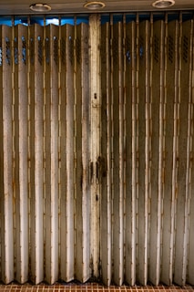 A weathered folding metal gate with vertical slats, exhibiting rust and grime. The gate is situated at the entrance of a structure with beige tiles on the ground. Three round lights are embedded in the ceiling above.