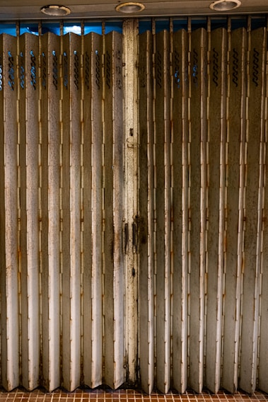 A weathered folding metal gate with vertical slats, exhibiting rust and grime. The gate is situated at the entrance of a structure with beige tiles on the ground. Three round lights are embedded in the ceiling above.