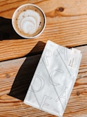 A cup of frothy cappuccino sits on a wooden table next to a package of coffee beans. The table has a natural wood grain and knots, adding a rustic feel. The coffee bag is designed with modern typography and a minimalistic style.