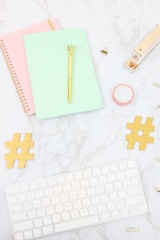 A desktop arrangement containing a mint green notebook with a gold pen on top, a pink spiral notebook, a gold stapler, a roll of pink washi tape, two gold hashtag symbols, a white keyboard, and several paper clips, all laid out on a marble surface.