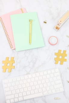 A desktop arrangement containing a mint green notebook with a gold pen on top, a pink spiral notebook, a gold stapler, a roll of pink washi tape, two gold hashtag symbols, a white keyboard, and several paper clips, all laid out on a marble surface.