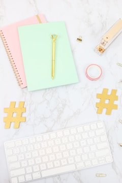 A desktop arrangement containing a mint green notebook with a gold pen on top, a pink spiral notebook, a gold stapler, a roll of pink washi tape, two gold hashtag symbols, a white keyboard, and several paper clips, all laid out on a marble surface.