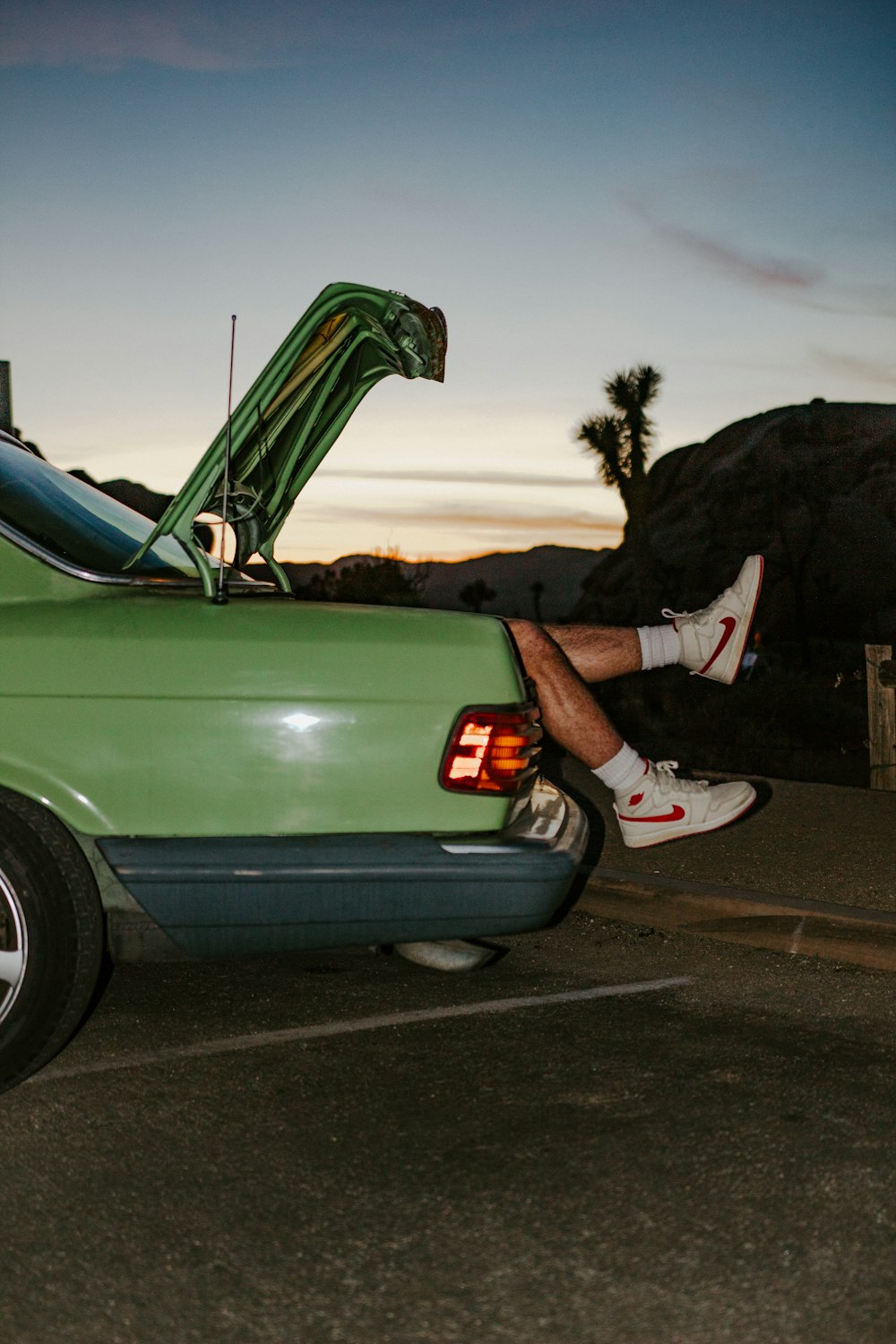 person in black and white nike sneakers sitting on green car hood during daytime