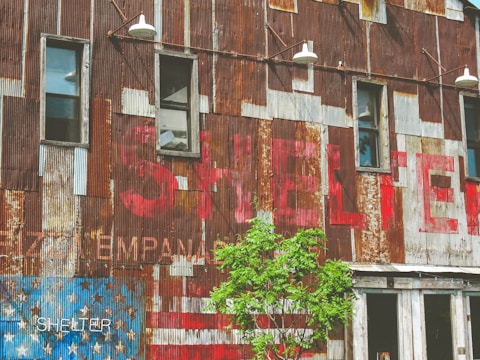A rustic building with corrugated metal siding, featuring faded red letters and an American flag design on the lower left. The building has several narrow windows and outdoor lights above them. A small green tree is in front of the building.