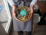 A person wearing a gray apron is holding a bowl filled with colorful ingredients, including greens, seeds, and possibly seafood or tofu, topped with a sprig of sprouts. The bowl has a lid with a label that reads 'my Bowl - POKE BAR-'. The person is standing indoors near a window, on a wooden floor.