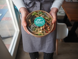 A person wearing a gray apron is holding a bowl filled with colorful ingredients, including greens, seeds, and possibly seafood or tofu, topped with a sprig of sprouts. The bowl has a lid with a label that reads 'my Bowl - POKE BAR-'. The person is standing indoors near a window, on a wooden floor.