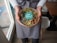 A person wearing a gray apron is holding a bowl filled with colorful ingredients, including greens, seeds, and possibly seafood or tofu, topped with a sprig of sprouts. The bowl has a lid with a label that reads 'my Bowl - POKE BAR-'. The person is standing indoors near a window, on a wooden floor.