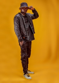 A person stands confidently in front of a plain mustard yellow background. They are wearing a dark quilted jacket, black pants, and a black hat. Their outfit is rounded off with light-colored sneakers and a visible chain necklace. The individual is holding the brim of their hat and looking towards the camera, exuding a sense of style and poise.