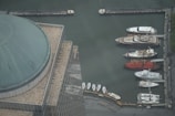 Aerial view of a marina with several luxury yachts docked along a quay. A large domed building is visible in the foreground, featuring a tiled roof with a grid pattern. The water appears calm, reflecting the industrial setting of the port.