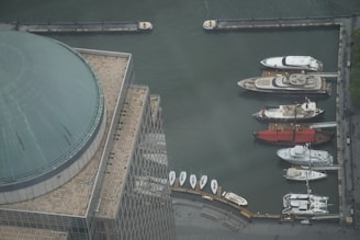 Aerial view of a marina with several luxury yachts docked along a quay. A large domed building is visible in the foreground, featuring a tiled roof with a grid pattern. The water appears calm, reflecting the industrial setting of the port.