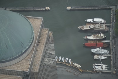 Aerial view of a marina with several luxury yachts docked along a quay. A large domed building is visible in the foreground, featuring a tiled roof with a grid pattern. The water appears calm, reflecting the industrial setting of the port.