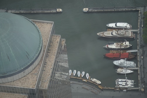 Aerial view of a marina with several luxury yachts docked along a quay. A large domed building is visible in the foreground, featuring a tiled roof with a grid pattern. The water appears calm, reflecting the industrial setting of the port.