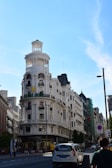 An elegant, historic building with a round turret and multiple balconies dominates the street. A Rolex sign is visible on the facade, indicating luxury retail. The street is lined with various buildings and a few pedestrians and vehicles are seen.