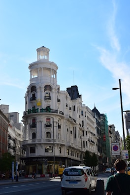 An elegant, historic building with a round turret and multiple balconies dominates the street. A Rolex sign is visible on the facade, indicating luxury retail. The street is lined with various buildings and a few pedestrians and vehicles are seen.