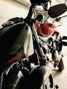 Two motorcycles are parked in an indoor setting. One bike, predominantly black, prominently features the brand name on the fuel tank. The other motorcycle appears to be red. The setting suggests an industrial or garage-like environment with pipes visible in the background.