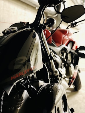 Two motorcycles are parked in an indoor setting. One bike, predominantly black, prominently features the brand name on the fuel tank. The other motorcycle appears to be red. The setting suggests an industrial or garage-like environment with pipes visible in the background.