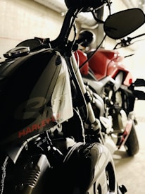 Two motorcycles are parked in an indoor setting. One bike, predominantly black, prominently features the brand name on the fuel tank. The other motorcycle appears to be red. The setting suggests an industrial or garage-like environment with pipes visible in the background.