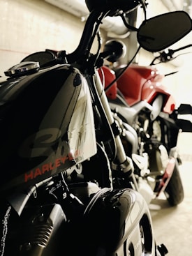Two motorcycles are parked in an indoor setting. One bike, predominantly black, prominently features the brand name on the fuel tank. The other motorcycle appears to be red. The setting suggests an industrial or garage-like environment with pipes visible in the background.