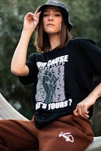 A person is wearing a black graphic t-shirt and brown pants, accessorized with a black bucket hat and large hoop earrings. The t-shirt has bold white text and a list of names.