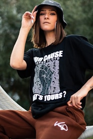 A person is wearing a black graphic t-shirt and brown pants, accessorized with a black bucket hat and large hoop earrings. The t-shirt has bold white text and a list of names.