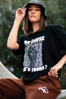 A person is wearing a black graphic t-shirt and brown pants, accessorized with a black bucket hat and large hoop earrings. The t-shirt has bold white text and a list of names.