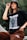 A person is wearing a black graphic t-shirt and brown pants, accessorized with a black bucket hat and large hoop earrings. The t-shirt has bold white text and a list of names.