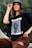 A person is wearing a black graphic t-shirt and brown pants, accessorized with a black bucket hat and large hoop earrings. The t-shirt has bold white text and a list of names.