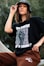 A person is wearing a black graphic t-shirt and brown pants, accessorized with a black bucket hat and large hoop earrings. The t-shirt has bold white text and a list of names.