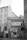 A black and white photograph of an urban street scene featuring an old building with a passageway labeled 'Na Zbořenci.' Above the archway are windows and a small balcony with flower boxes. A parked car is partially visible through the archway, and two people stand near the entrance. The cobblestone street, graffiti on walls, and several signs add to the city atmosphere.
