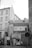 A black and white photograph of an urban street scene featuring an old building with a passageway labeled 'Na Zbořenci.' Above the archway are windows and a small balcony with flower boxes. A parked car is partially visible through the archway, and two people stand near the entrance. The cobblestone street, graffiti on walls, and several signs add to the city atmosphere.