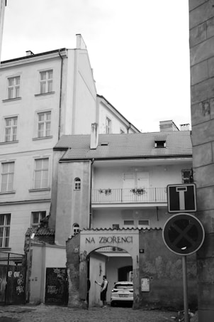 A black and white photograph of an urban street scene featuring an old building with a passageway labeled 'Na Zbořenci.' Above the archway are windows and a small balcony with flower boxes. A parked car is partially visible through the archway, and two people stand near the entrance. The cobblestone street, graffiti on walls, and several signs add to the city atmosphere.