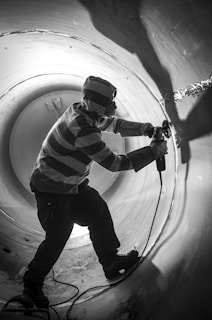 A person wearing a striped hoodie and protective goggles is using a power tool inside a large cylindrical metal structure. The lighting creates dramatic shadows, highlighting the contours and texture of the surface.