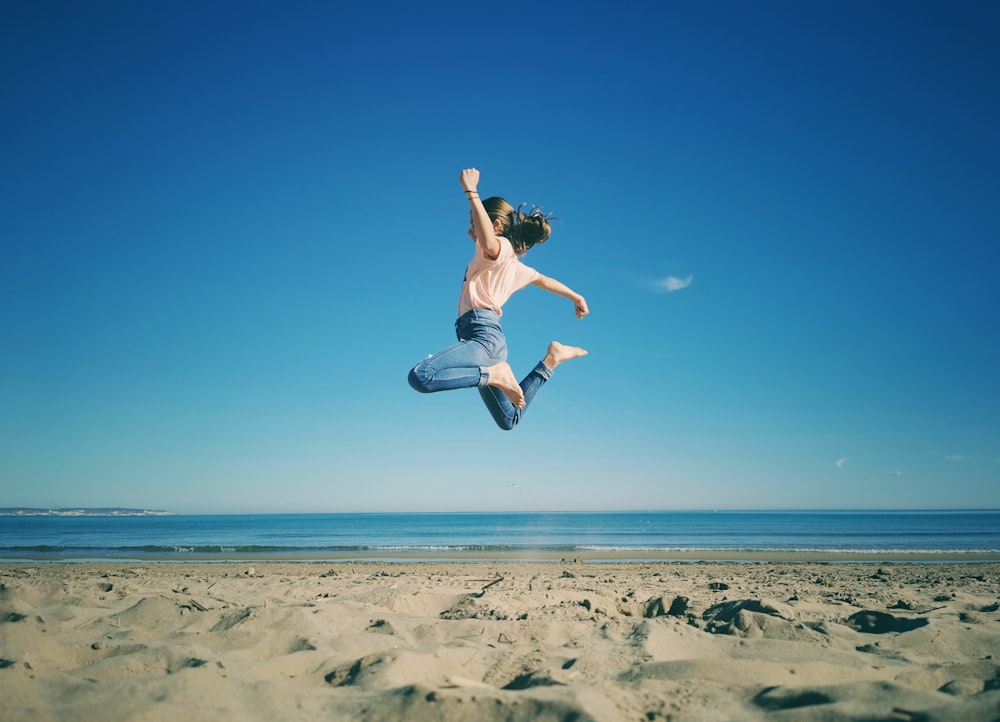 昼間にビーチでジャンプする白いタンクトップと青いデニムのショート パンツの女性